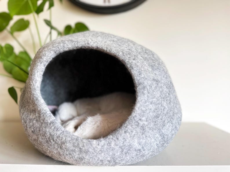 May include: A rounded, light gray felt pet cave with a large, circular opening. Inside the dark interior of the structure, a small, white, furry animal is curled up and sleeping, with its soft white paws visible near the front edge of the opening.
