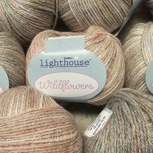 May include: A close-up of several skeins of yarn in a beige and pink colorway. The yarn is labeled "Lighthouse Wildflowers".