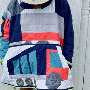 Puede incluir: Un suéter acolchado con capucha que presenta un diseño de patchwork con un gráfico grande de un camión volquete en rojo, azul y gris. La prenda incluye mangas largas, un borde de dobladillo verde claro y un sombrero de paja con una banda de cuero marrón.
