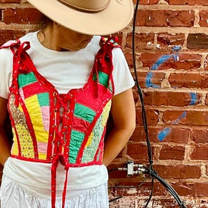 May include: A colorful patchwork corset with red lacing, paired with a white top and skirt. The outfit is accessorized with a tan hat and light green sandals. The background is a red brick wall.