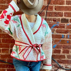May include: A cream-colored quilted jacket with red and green floral patterns and a red tie belt. The jacket is paired with blue jeans, a tan hat, and green boots. The background is a red brick wall.