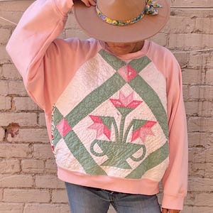 May include: A light pink sweatshirt featuring a central quilted panel with a green and white geometric pattern and a pink floral motif. The outfit includes a tan fedora hat with a floral band, blue denim jeans, white socks, and red leather shoes.