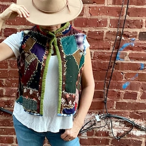 May include: A woman wearing a brown hat, a white t-shirt, blue jeans, and a multi-coloured patchwork vest with a green scarf tied around the neck.