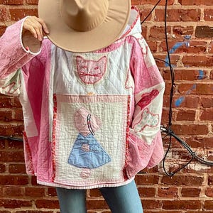 Puede incluir: Chaqueta acolchada rosa y blanca con capucha y aplicación de gato. La chaqueta presenta un diseño de patchwork con un panel azul y blanco. La persona lleva vaqueros azules, sombrero beige y botas verdes.