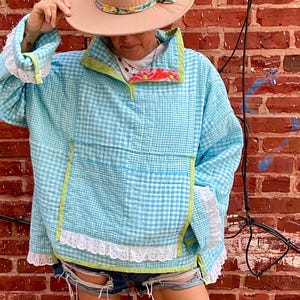 May include: A light blue and white gingham quilted pullover with a high collar, lime green trim, and white eyelet lace trim on the cuffs and hem. The outfit includes a tan wide-brimmed hat with a floral band and light blue leather cowboy boots.