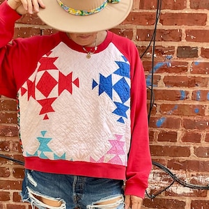 Puede incluir: Un sombrero fedora beige con una banda floral, una sudadera roja con un panel frontal acolchado blanco con patrones geométricos en rojo, azul, rosa y turquesa, pantalones cortos de mezclilla azul desgastados y botas vaqueras de cuero azul claro.