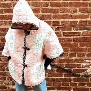 Op de afbeelding: Een roze gewatteerd jack met capuchon met een zwart-witte rand. Het jack heeft een knoopsluiting en een patroon van kleine dieren. De persoon die het jack draagt, staat voor een bakstenen muur en draagt zwarte laarzen.