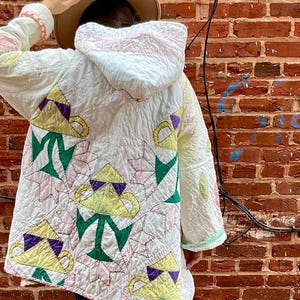 May include: A quilted white coat with a hood, featuring a repeating pattern of yellow teacups with purple accents and green leaves. The coat is worn with blue jeans and tan boots. The background is a brick wall.