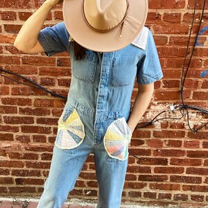 Può includere: Una persona indossa una tuta di jeans con tasche a patchwork e un cappello fedora marrone. La tuta ha una chiusura con bottoni sul davanti e maniche corte.