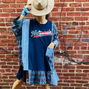 Op de afbeelding: Een patchwork jurk met een marineblauwe top met het opschrift "Nationals", gecombineerd met geruite en gestreepte panelen. De jurk heeft een gefranjerde zoom en lange mouwen. Een beige hoed en sandalen maken de look compleet.