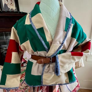May include: A patchwork robe in green, red, and cream colors, with a brown leather belt. The robe is displayed on a mannequin, showcasing its quilted design and unique color block pattern. The robe's sleeves are wide and the collar is trimmed with blue fabric.