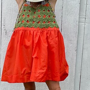 May include: A sleeveless dress featuring a bright orange skirt and a green crocheted bodice with a floral pattern. The dress is paired with a light-colored straw hat with a brown band and metallic silver thong sandals with a buckle closure.