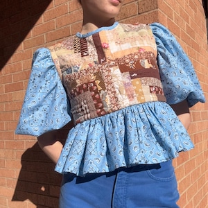 May include: A patchwork top with a blue paisley print and a ruffled hem. The bodice features a variety of fabric patterns in shades of brown, beige, and red. The sleeves are a light blue paisley print. The top is paired with blue jeans.