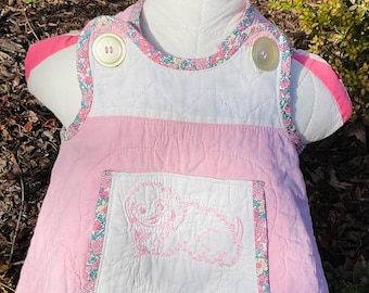 May include: A pink quilted baby dress with a white embroidered puppy on the front pocket. The dress has a floral trim and two white buttons on the straps.