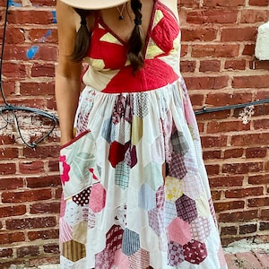 May include: A woman wearing a white dress with a red and yellow patchwork design. The dress has a hexagonal pattern and a large pocket on the side. She is wearing a brown hat and brown sandals.