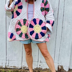 May include: A quilted jacket featuring a white base with large, circular floral patterns in shades of pink, dark blue, and yellow. The jacket has long sleeves, a rounded neckline, and a white trim. A light-colored straw hat is worn with the jacket.