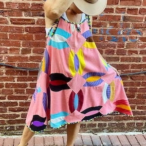May include: A pink patchwork dress with colorful geometric patterns, including circles and squares. The dress has a black and yellow trim and is paired with yellow sandals. A beige hat is held up.
