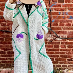 May include: A long, quilted coat in white with green trim and purple floral appliqués. The coat features a diamond pattern and is worn with a tan hat. The coat is a unique, handmade piece.
