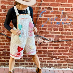 Può includere: Salopette corta trapuntata color crema con toppe applicate colorate. La salopette presenta un design floreale rosso, rosa e verde. La modella indossa un cappello fedora beige e stivaletti marroni. Lo sfondo è un muro di mattoni rossi.