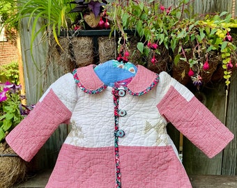 May include: A white quilted jacket with red and white gingham trim. The jacket has a white collar with a red and white gingham trim and a row of buttons down the front. The jacket is quilted with a white fabric and has a red and white gingham pattern on the bottom.