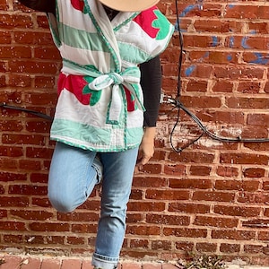 May include: A quilted vest with a red and green strawberry design, tied at the waist. The vest is worn over a black long-sleeve shirt and paired with light blue jeans and light green boots. A tan fedora hat is worn.