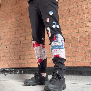 May include: Black sweatpants with colorful patchwork details on the lower legs. The patchwork includes red, white, blue, and pink fabric pieces. The pants are paired with black sneakers and a black sweatshirt.