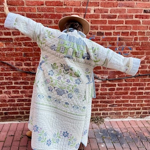 May include: A long, quilted coat in cream with blue and green floral and house motifs. The coat features a detailed pattern of flowers, vases, and small houses. The coat has a relaxed fit and is worn open, showcasing the intricate design.