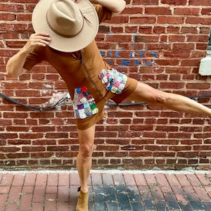 May include: A brown romper with a front zipper and patchwork details. The outfit is paired with a tan fedora hat and matching peep-toe booties. The background features a brick wall and a brick-paved walkway.