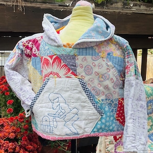 Peut inclure: Un sweat à capuche patchwork avec divers motifs floraux et géométriques dans des tons de bleu, rouge et blanc. La poche avant présente un motif brodé bleu. Le sweat à capuche est présenté sur un mannequin.