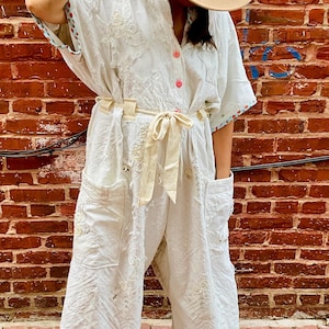 May include: A white jumpsuit with short sleeves, a button-up front, and a matching belt. The jumpsuit features embroidered details and pockets. A beige hat is worn. The background is a red brick wall.