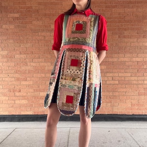 May include: A patchwork dress with a red square design, worn over a red long-sleeve shirt. The dress features a variety of fabric patterns and colors, including pink, blue, and green. The dress has a pleated skirt and is worn with black sneakers.