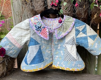 May include: A patchwork baby jacket in a purple, white and blue colour scheme. The jacket has a ruffled collar and is made of quilted fabric.