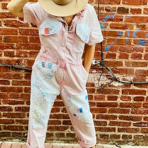 May include: A light pink jumpsuit with patchwork details on the chest and legs. The short-sleeved jumpsuit has a button-up front and is paired with a tan hat and yellow sandals. The background is a red brick wall.