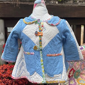 May include: A handmade, quilted baby jacket in blue, white, and floral patterns. The jacket features a Peter Pan collar, two front pockets, and decorative button closures. The jacket is displayed on a mannequin.