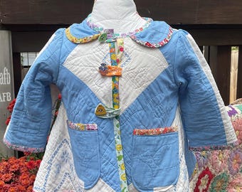 May include: A handmade, quilted baby jacket in blue, white, and floral patterns. The jacket features a Peter Pan collar, two front pockets, and decorative button closures. The jacket is displayed on a mannequin.