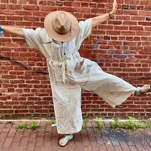 Op de afbeelding: Een crèmekleurige jumpsuit met kleurrijke geborduurde details en een bijpassende riem. De outfit wordt gecombineerd met een bruine fedora hoed en lichtblauwe sandalen. De achtergrond is een rode bakstenen muur.