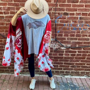 Può includere: Una maglietta grigia con una grafica Razorback è indossata sotto un poncho patchwork rosso e bianco. Viene indossato un cappello color camoscio con una fascia marrone. La persona indossa jeans blu scuro e scarpe da ginnastica bianche. Lo sfondo è un muro di mattoni rossi.