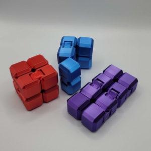 Puede incluir: Tres cubos anti estrés de diferentes colores: rojo, azul y morado. Cada cubo tiene múltiples lados con diferentes texturas y formas.