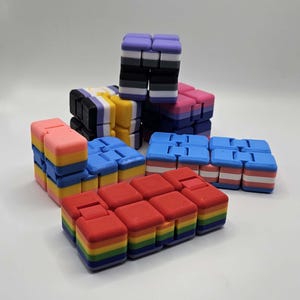 May include: A set of six colorful fidget cubes, each with a different pattern. The cubes are made of silicone and have a smooth, tactile surface. The colors include pink, yellow, blue, black, white, and red.
