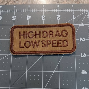 May include: A brown embroidered patch with a white outline. The patch reads "HIGH DRAG LOW SPEED".