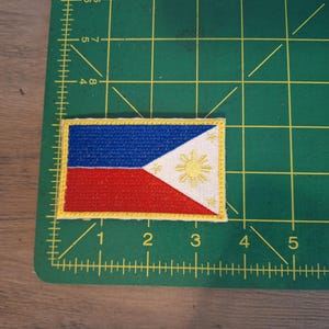 May include: A yellow embroidered patch of the Philippine flag. The flag is red, white, and blue with a yellow sun and stars.
