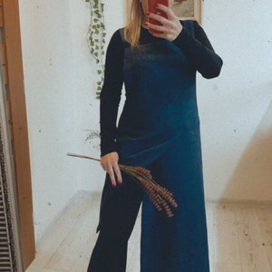 Może przedstawiać: Kobieta w długiej, ciemnoniebieskiej sukience z długim, czarnym rękawem pod spodem. Sukienka ma design w stylu otulającego i rozcięcie z boku. Trzyma bukiet suszonych kwiatów.