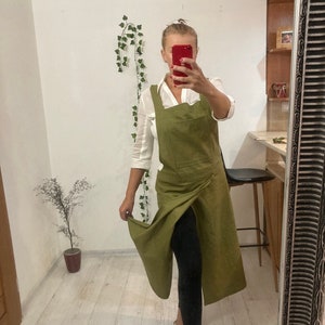 Może przedstawiać: Długi, oliwkowo-zielony fartuch lniany w stylu wiązanym z jedną kieszenią. Fartuch jest noszony na białej koszuli i czarnych legginsach.