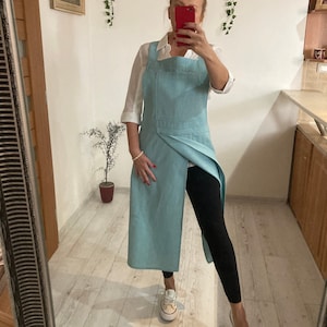 Może przedstawiać: Jasnoniebieski fartuch o unikalnym kroju. Fartuch ma dużą przednią kieszeń i długą, zwiewną spódnicę. Fartuch jest wykonany z teksturowanej tkaniny i jest noszony na białej koszuli i czarnych legginsach. Stylowy dodatek do każdej kuchni.