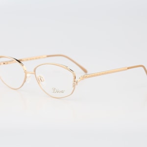 Puede incluir: Gafas de metal en tono dorado con montura de forma ovalada. Las varillas presentan un patrón de cuentas texturizado en los laterales. El cristal derecho tiene grabado el nombre Diva en letra cursiva. El puente y las bisagras son finos.