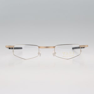 May include: Gold-tone metal frame glasses with a geometric shape. The glasses have a thin metal frame and clear lenses. The brand name "TAXI" is printed on the right lens.
