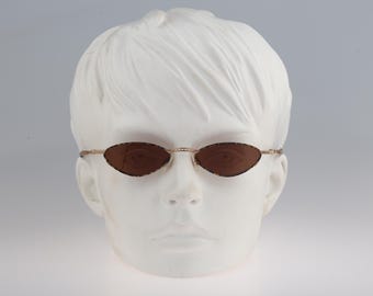 May include: A pair of oval-shaped sunglasses with a thin gold frame and brown lenses. The sunglasses are on a white mannequin head.