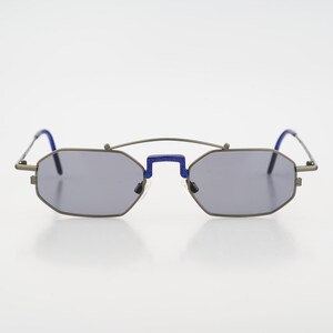 Puede incluir: Gafas de sol geométricas con lentes grises y montura de metal. El puente y las patillas presentan detalles azules. Las gafas tienen una forma angular única.