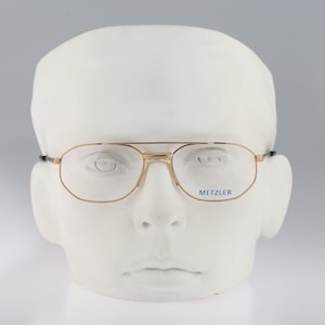 May include: Gold-rimmed glasses with clear lenses on a white mannequin head. The glasses have a blue "METZLER" logo on the right lens.