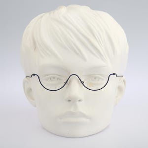Puede incluir: Un par de gafas de lectura de media luna negras con monturas de metal fino. Las gafas presentan una forma de lente semicircular distintiva y un diseño de puente alto, sobre una cabeza de maniquí blanca. Monturas minimalistas y finas.